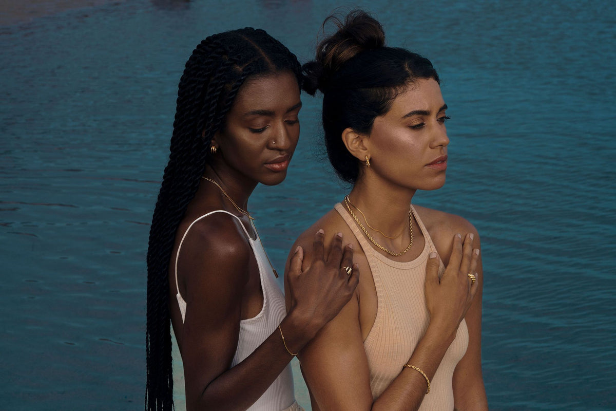 Two women standing close together by deep blue water, wearing layered gold-toned jewelry and neutral tops.