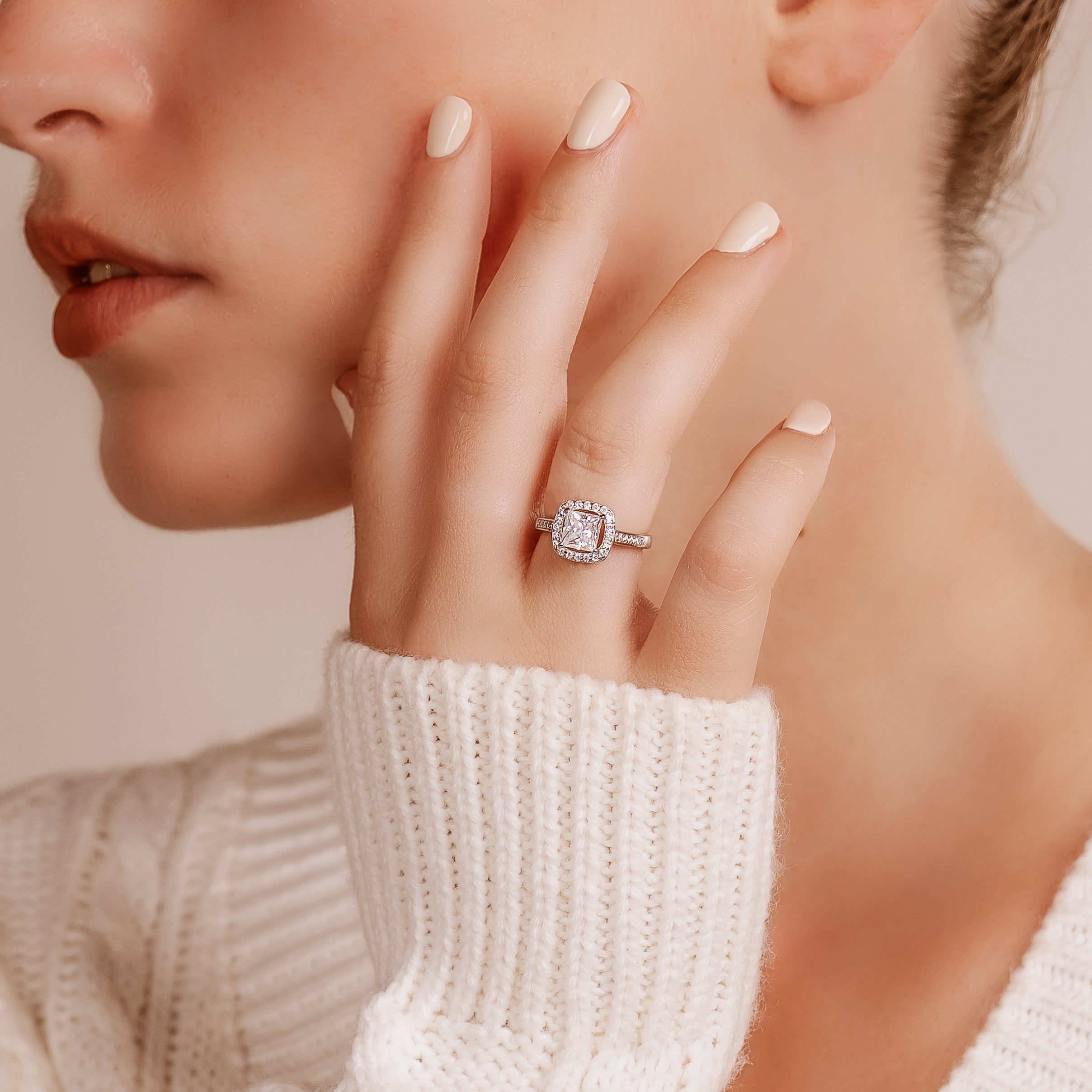 Close-up of a woman's hand resting on her cheek, showing a silver-toned halo ring with a square center stone
