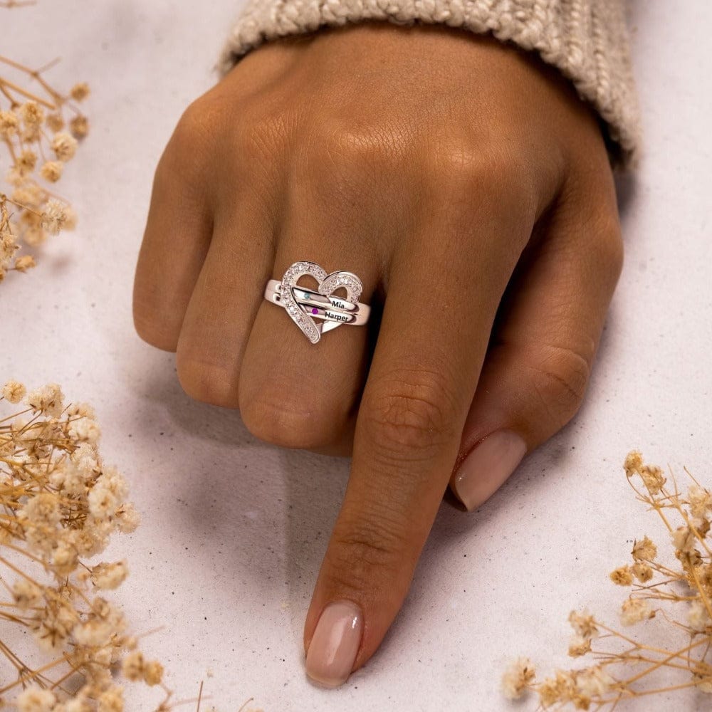 Hand wearing a silver-toned heart ring with two engraved name plates and small colored stones.