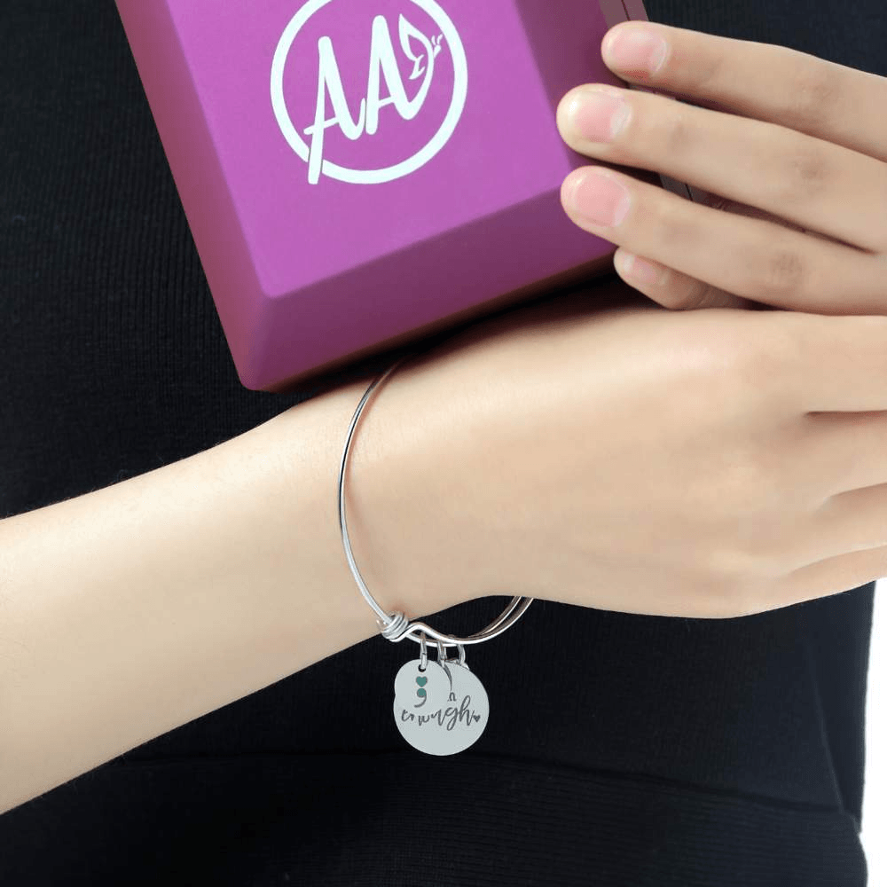 Silver-toned charm bangle with a round 'enough' pendant on a wrist, hand holding a purple jewelry box.