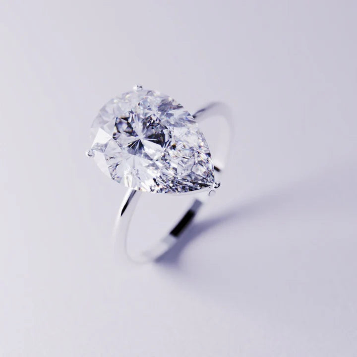 A silver-toned solitaire ring featuring a large clear pear-cut gemstone on a light background.