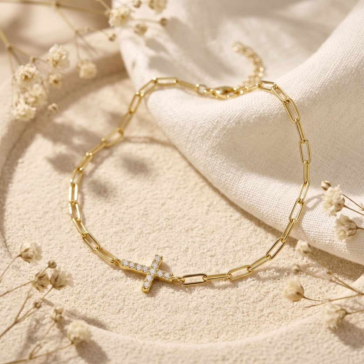 Gold-tone paperclip chain bracelet with a pave-set cross on a textured beige surface.