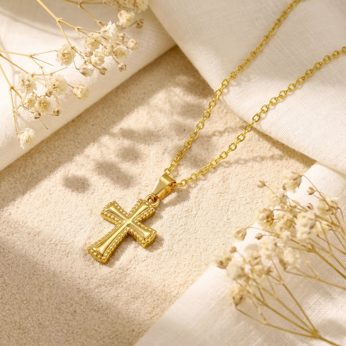 Gold-tone beaded cross pendant necklace flat lay on sand with cream linen fabric and dried flowers.