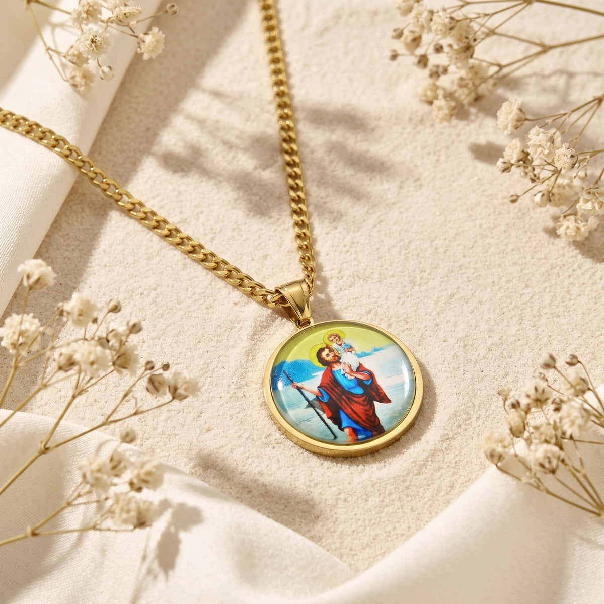Gold-tone St. Christopher medallion necklace lying flat on sand surrounded by white dried flowers and cream fabric.