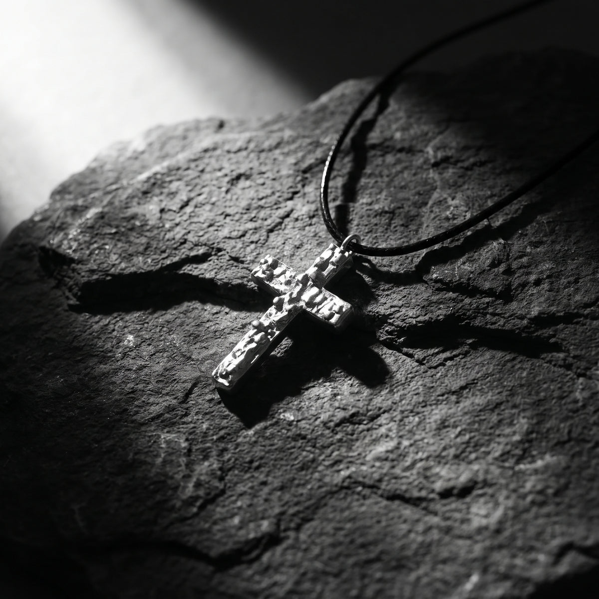 Textured silver cross pendant on a black cord lying on a rough dark rock surface.