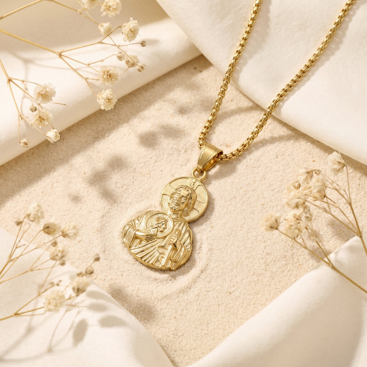 Gold finish sacred icon pendant lying on sand, framed by white fabric and dried flowers.