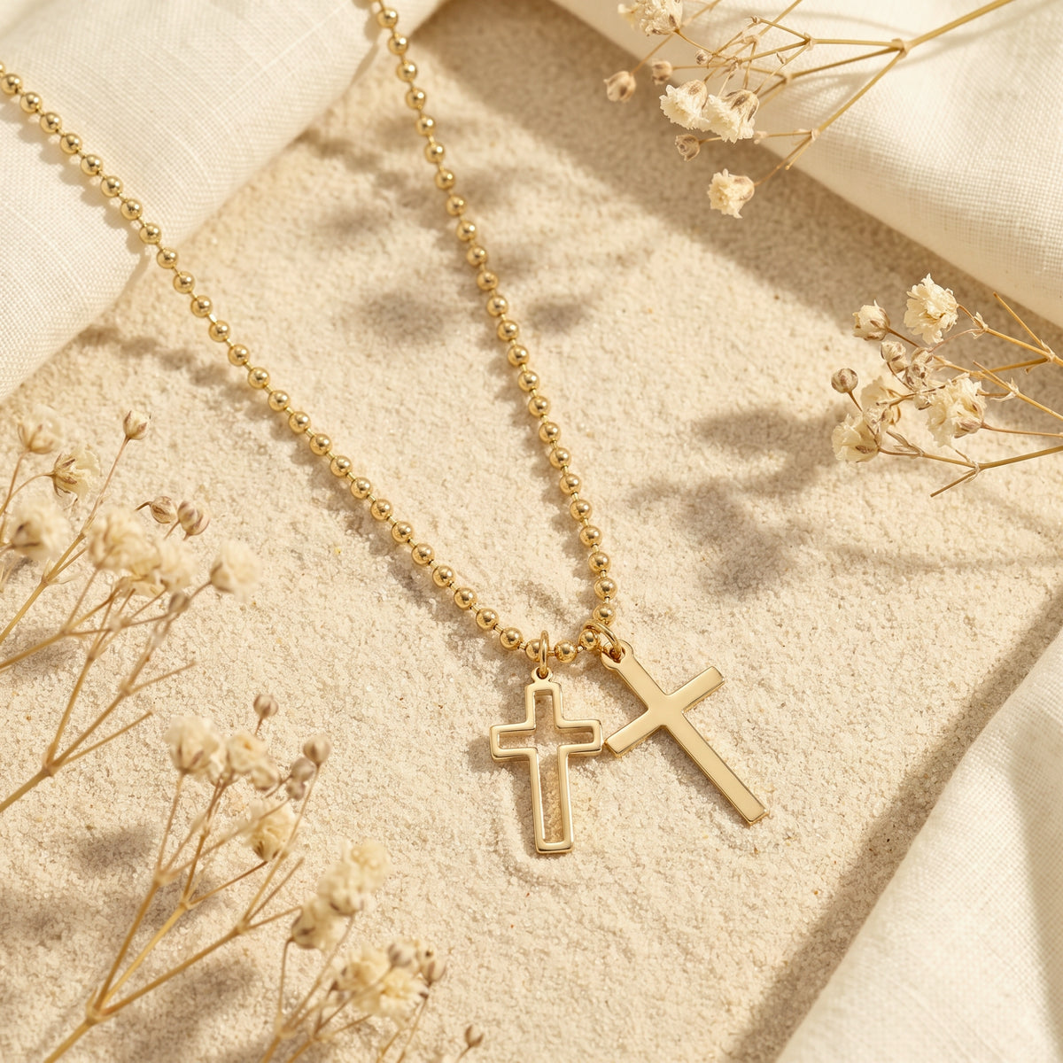 Gold-tone double cross necklace resting on fine sand with dried white flowers and cream linen.