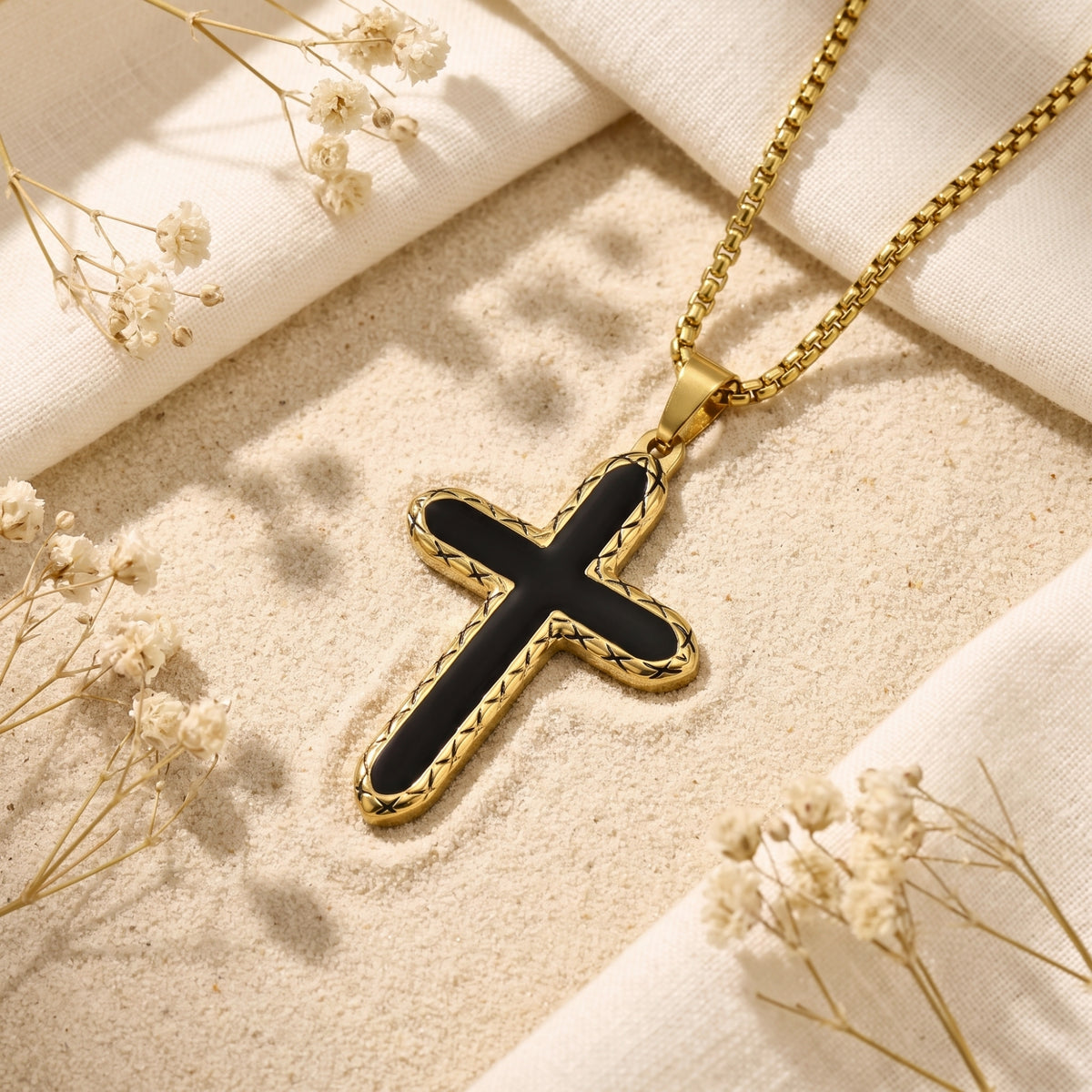 Gold-tone cross pendant with black inlay on sand beside white fabric and dried flowers.