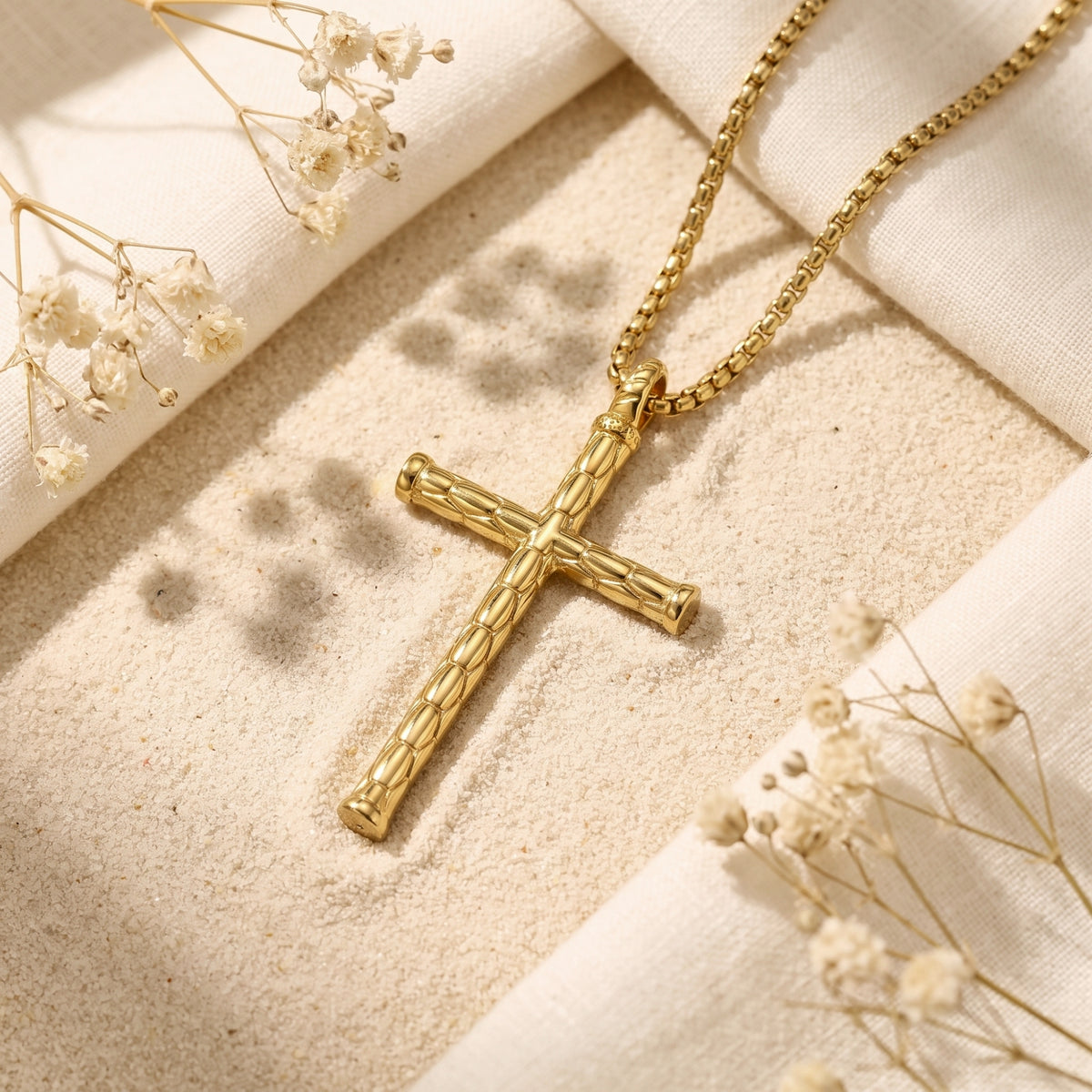 Gold-tone segmented cross necklace resting on sand beside white fabric and dried flowers.