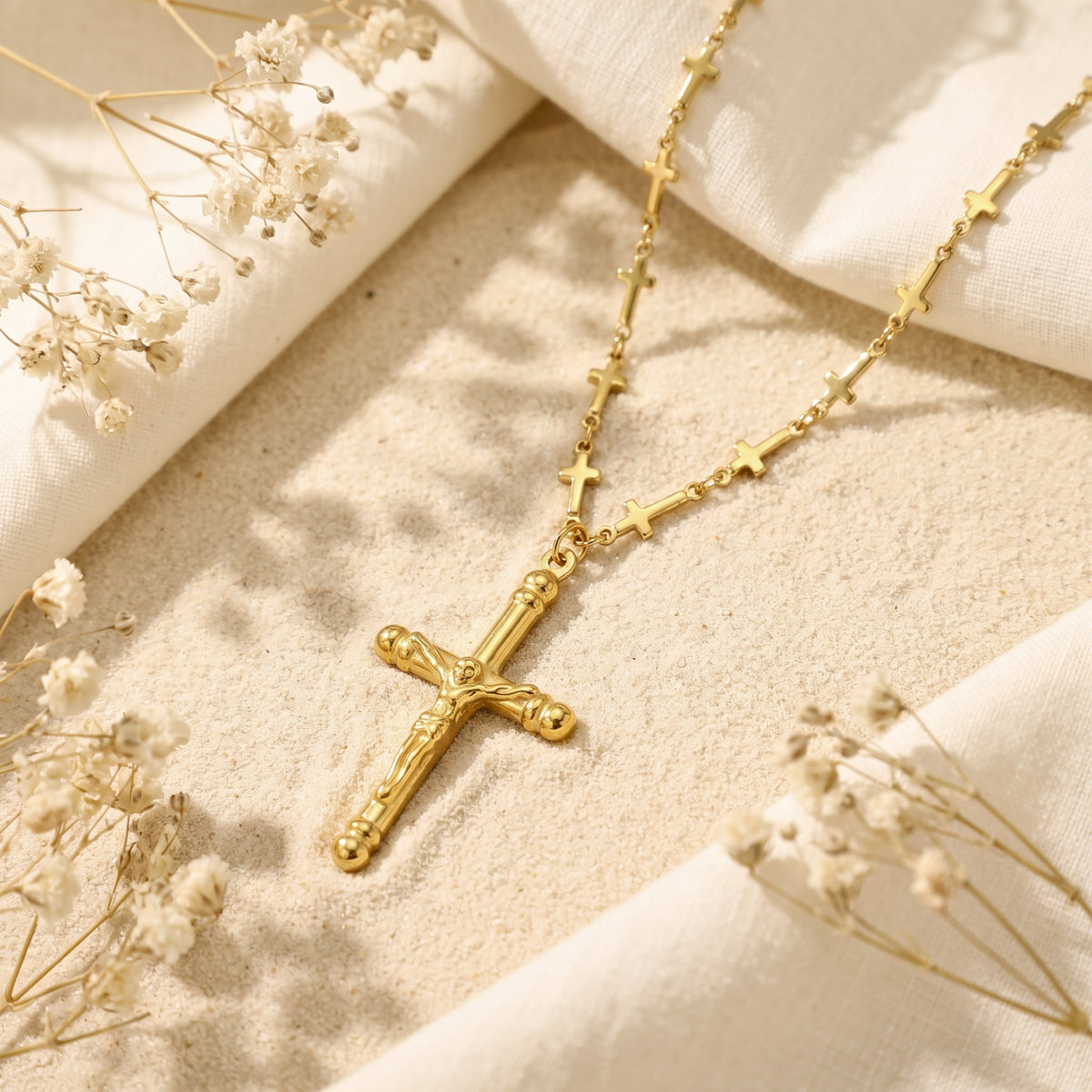 Gold-tone station crucifix necklace lying on fine sand beside white fabric and dried flowers.