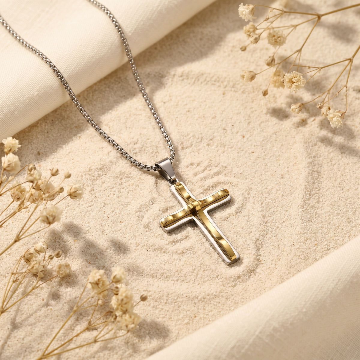 Two-tone cross necklace with a silver and gold-tone pendant lying on sand beside dried white flowers.