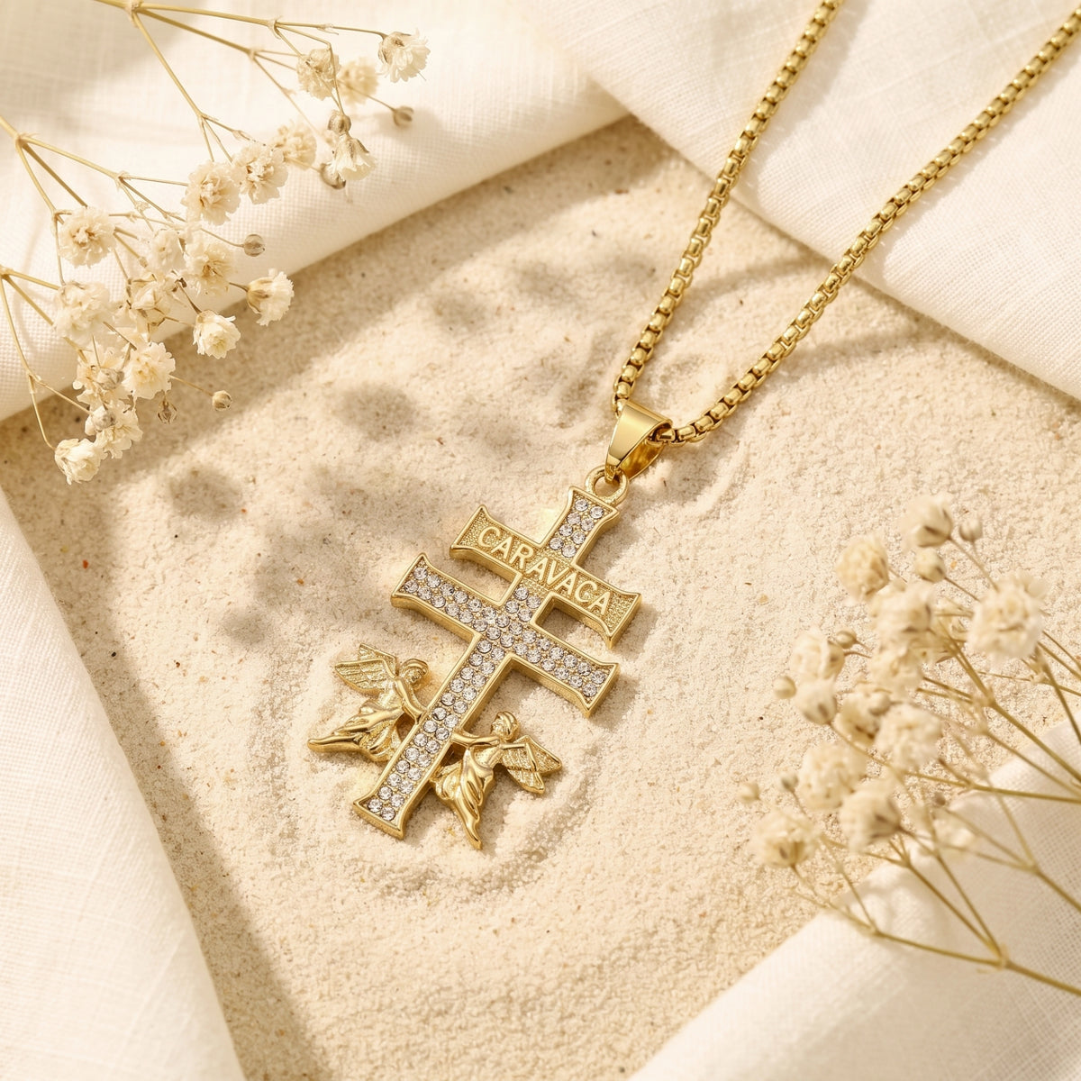 Gold Caravaca cross pendant with clear crystal accents laid flat on sand with dried flowers.