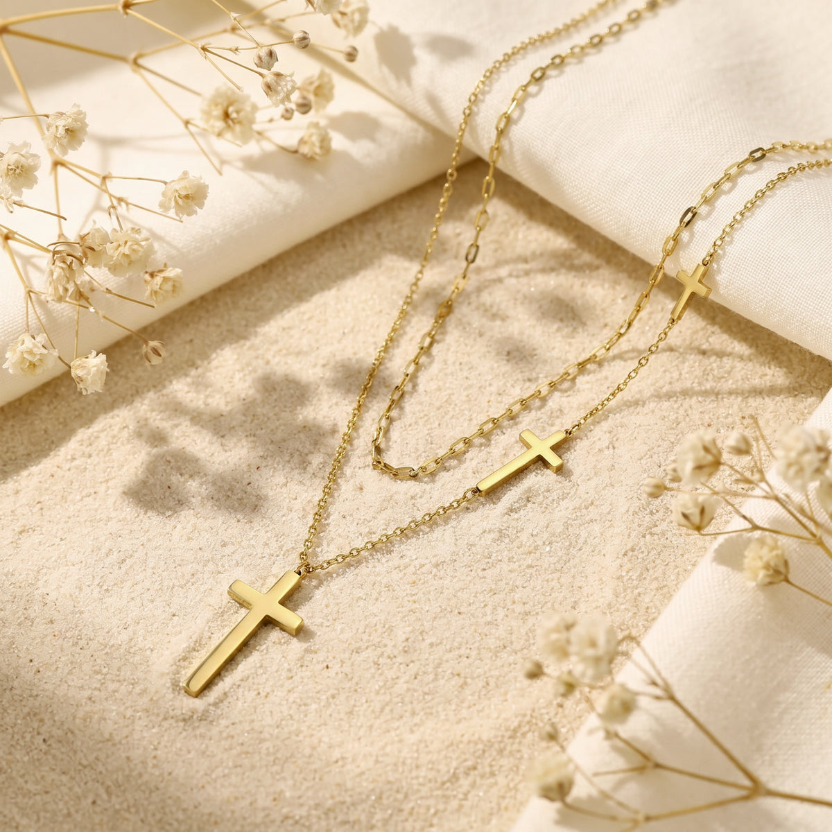Gold-tone layered cross necklace arranged diagonally on sand with folded white fabric and dried white flowers.