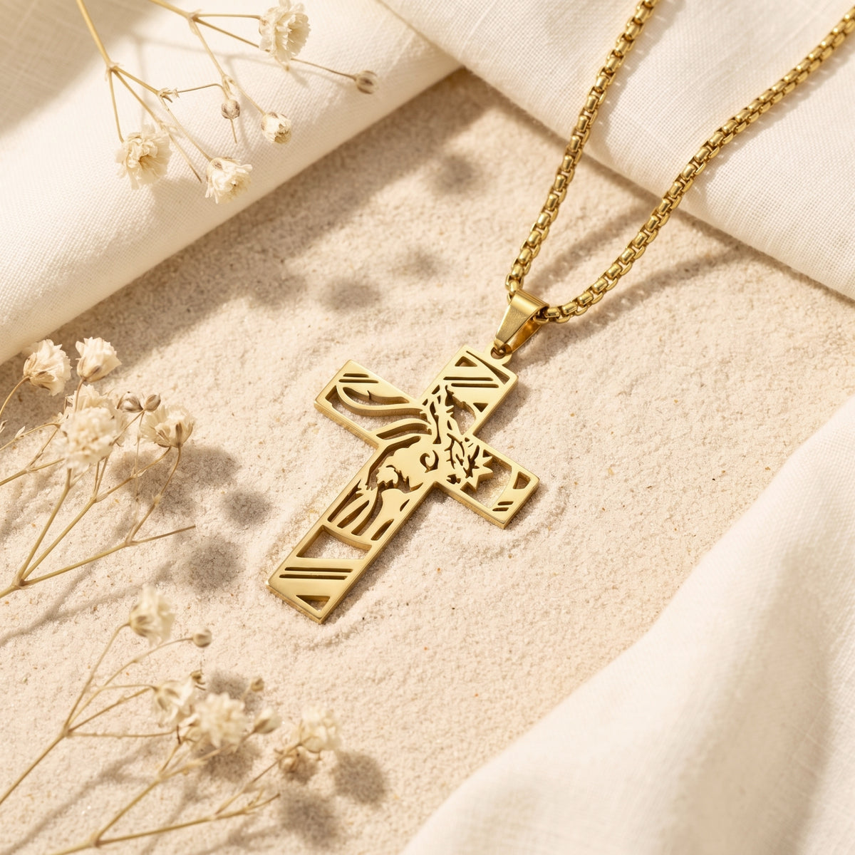 Flat lay of a gold-tone sculpted cross necklace on sand with white fabric and dried flowers.