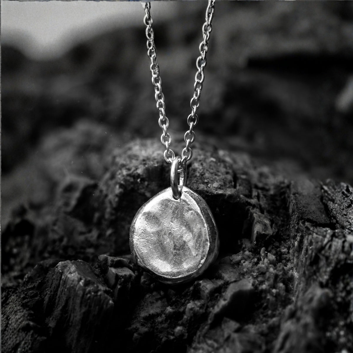 Close-up of a silver textured pebble pendant hanging from a silver chain over dark rock.