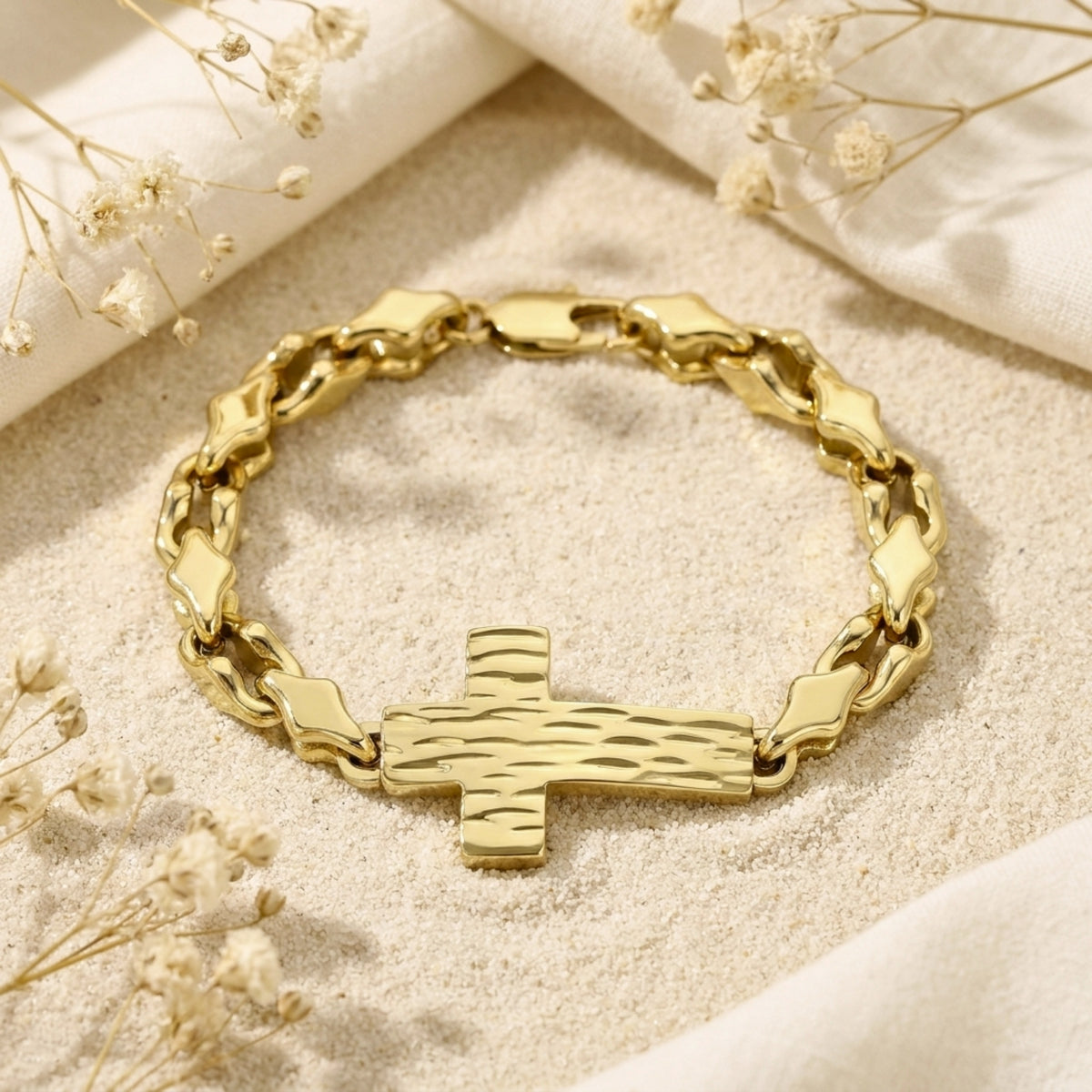 Gold-tone chain link bracelet with a textured cross centerpiece, displayed flat on sand among dried flowers.