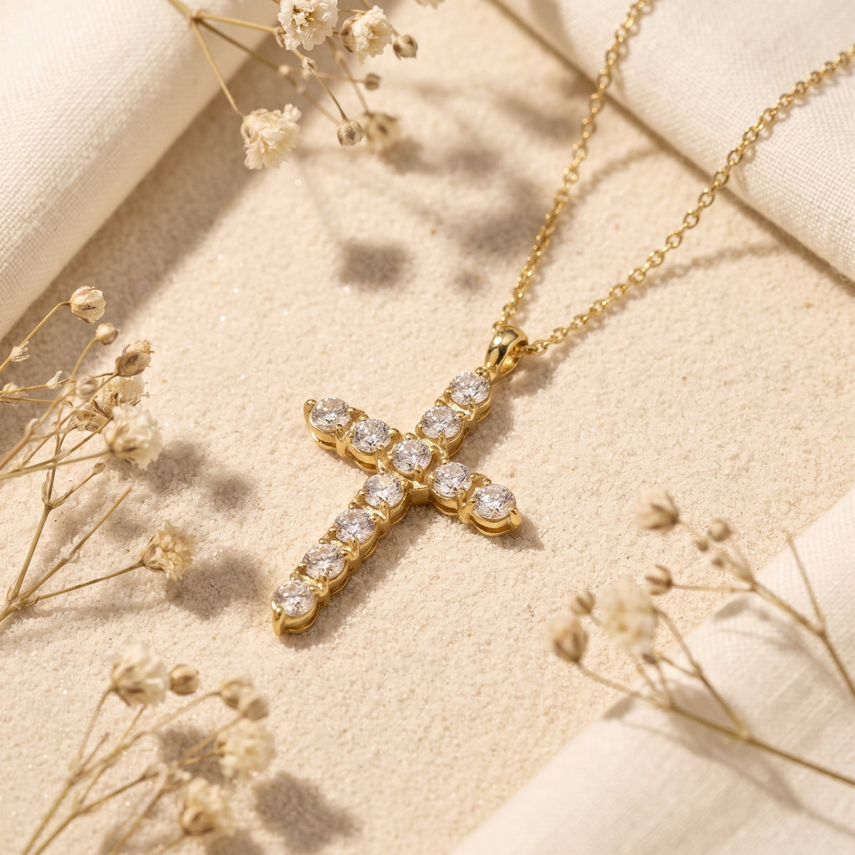 Gold-tone cross necklace with round clear stones in prong settings, resting on sand beside dried flowers.