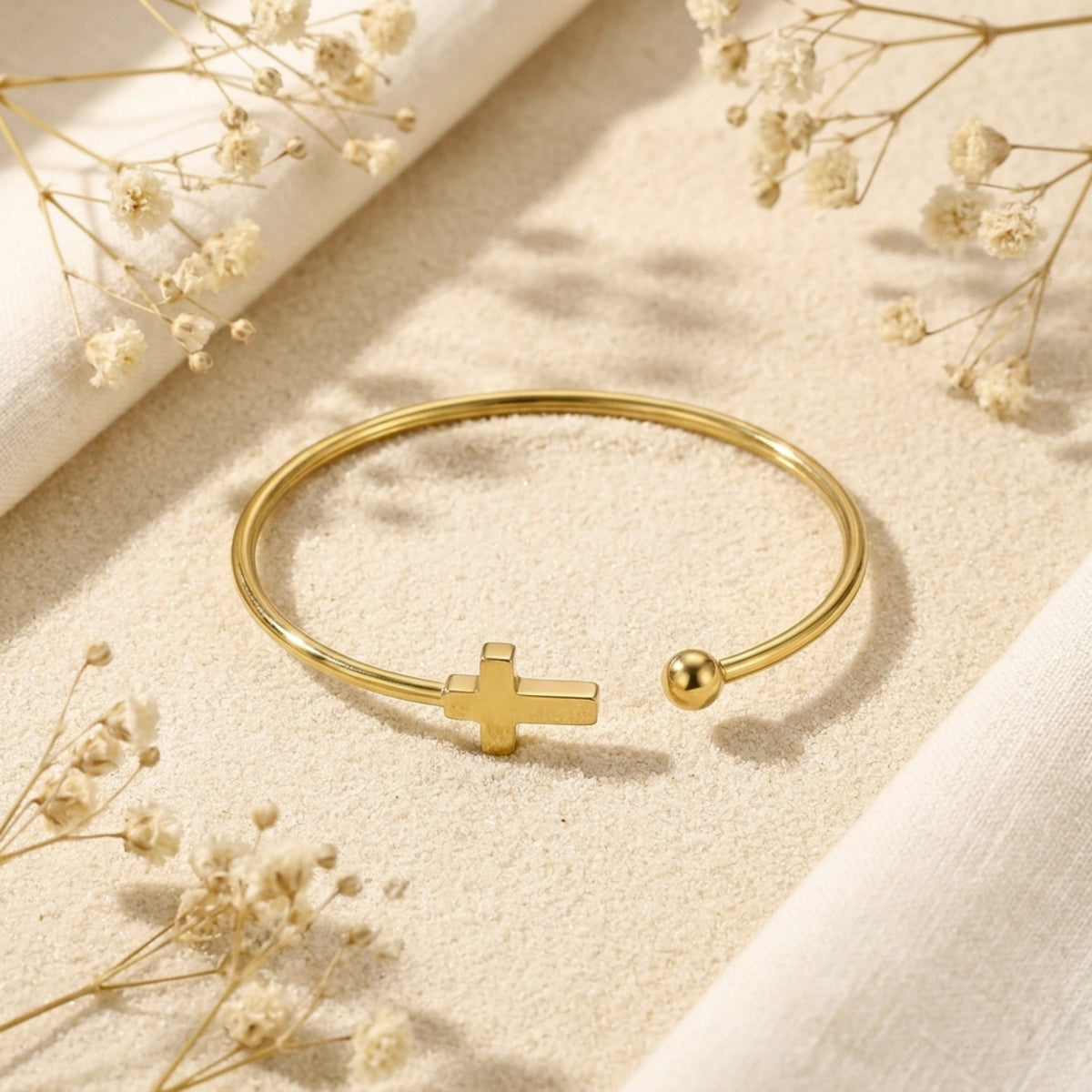 Gold-tone open cuff bracelet featuring a cross and ball end, resting flat on fine sand with white floral accents.