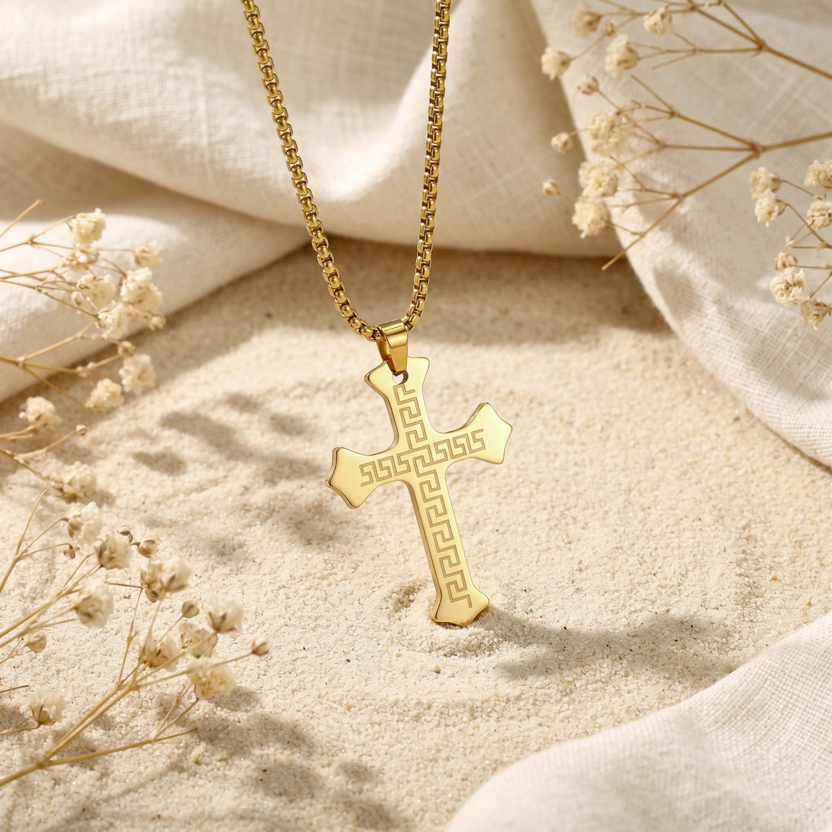 Gold-toned Greek key cross necklace arranged on sand and white fabric with dried flowers.