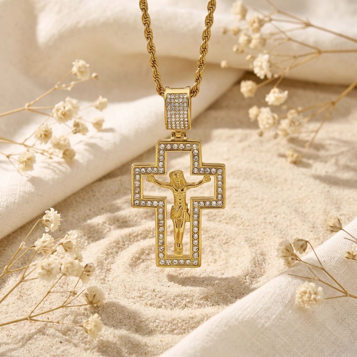 Gold-tone cutout crucifix pendant with clear stones hanging over sand among white dried flowers.
