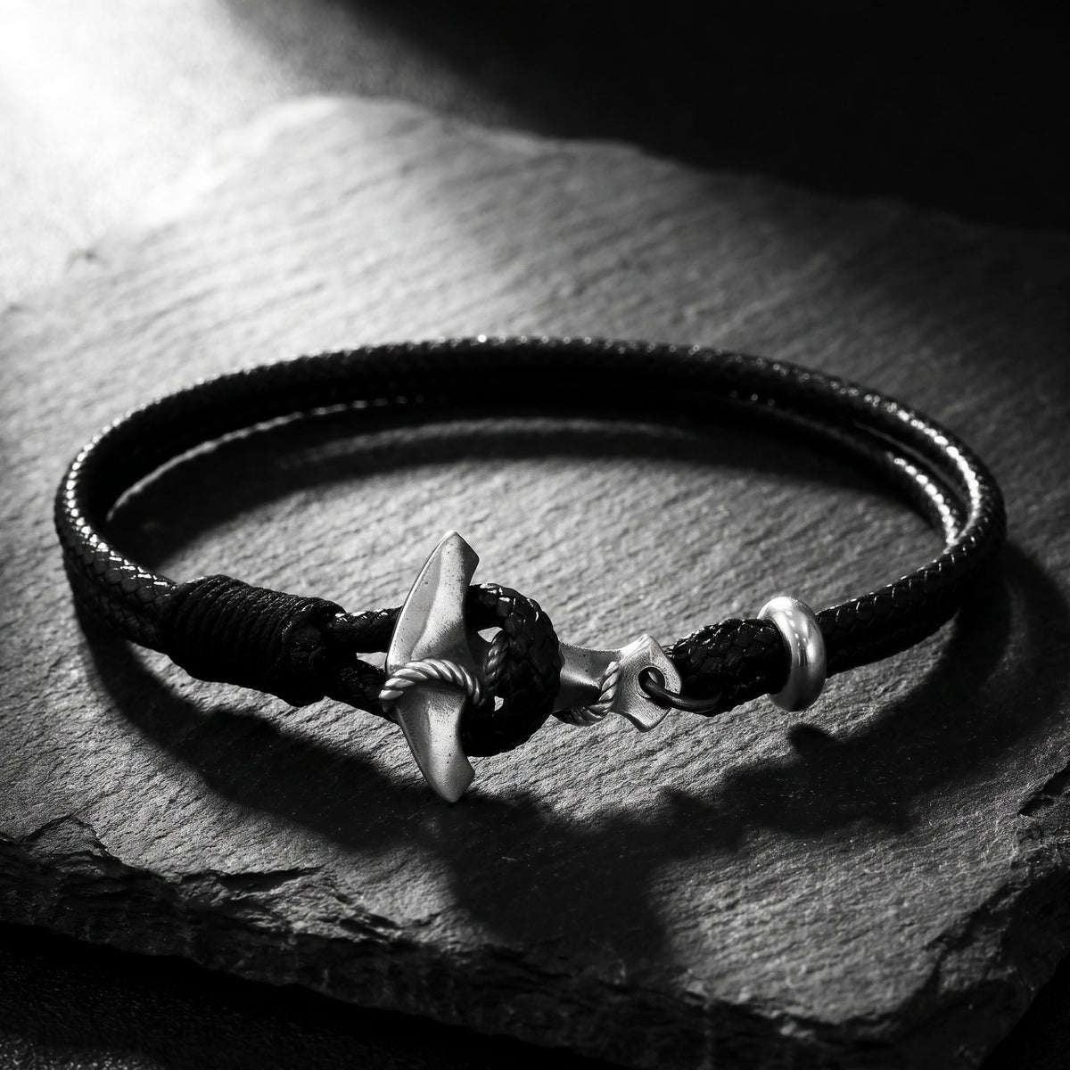 Black braided cord bracelet with a silver anchor toggle clasp on a dark slate surface.