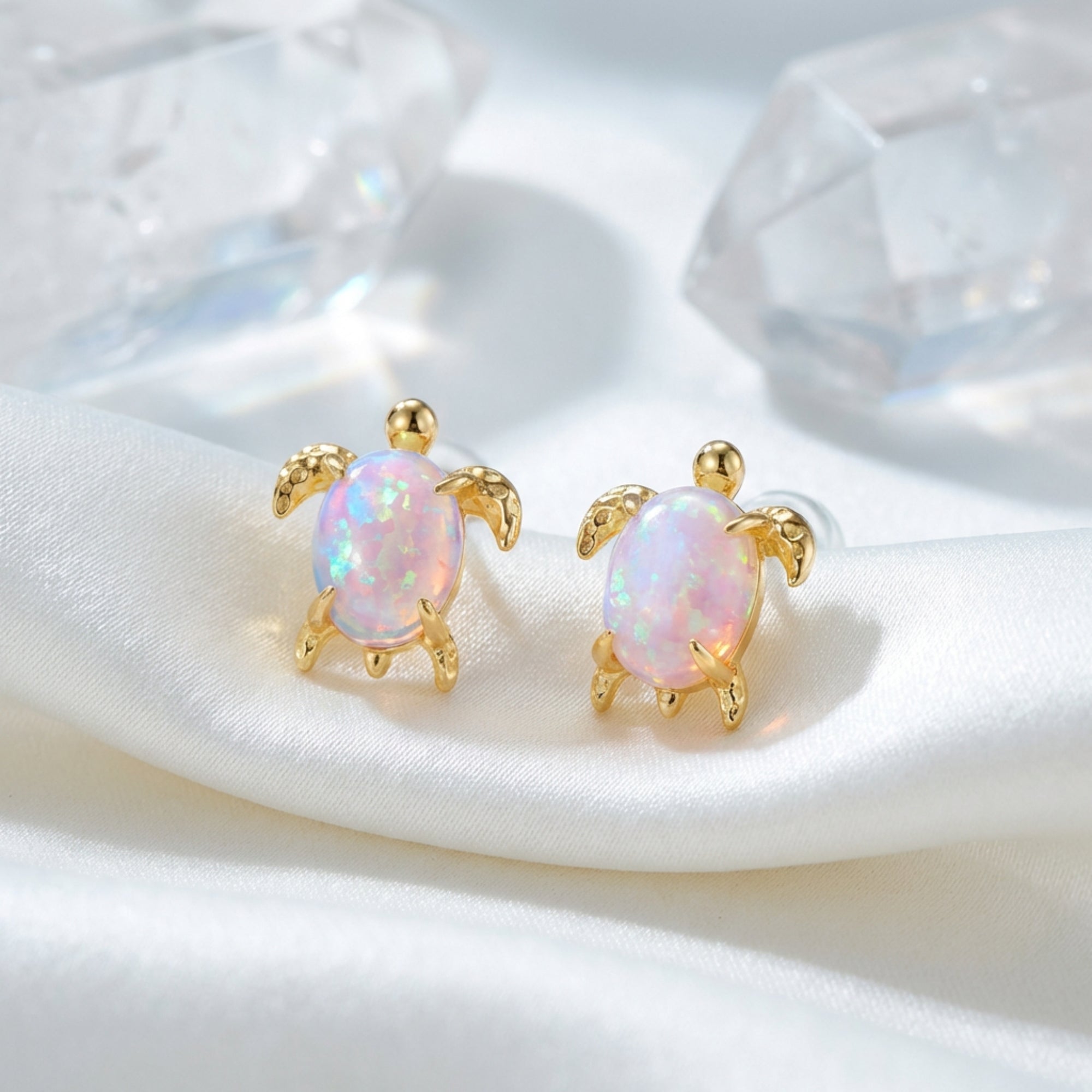 Gold-tone turtle stud earrings with oval white opals on white silk fabric with crystal accents.