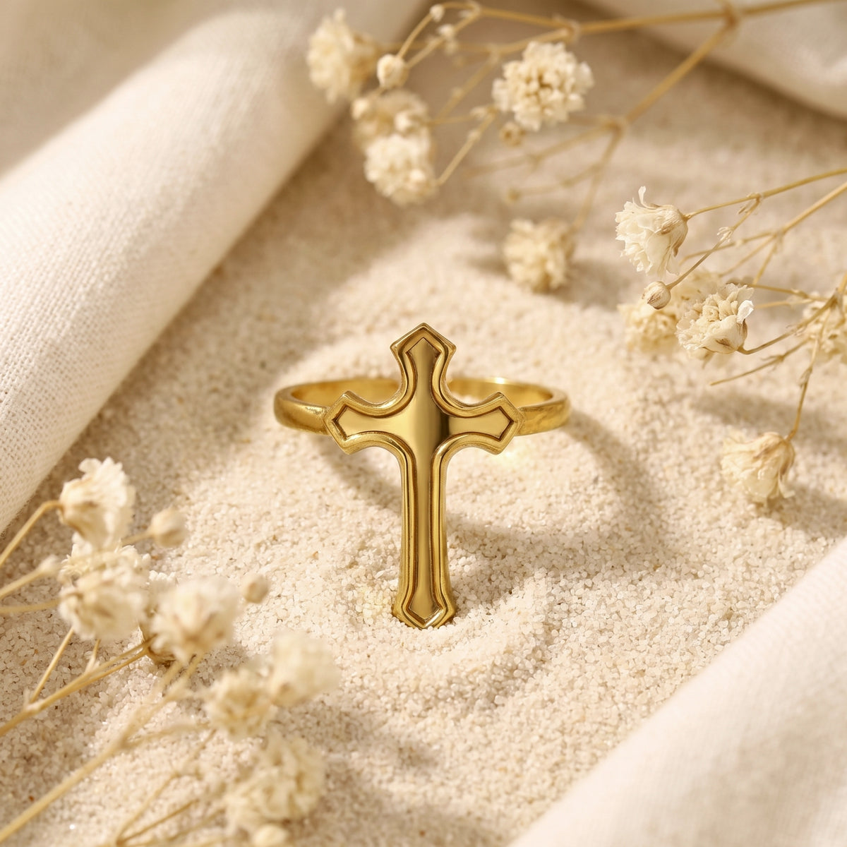 Frontal view of the gold-tone cross ring standing on fine white sand among dried flowers.
