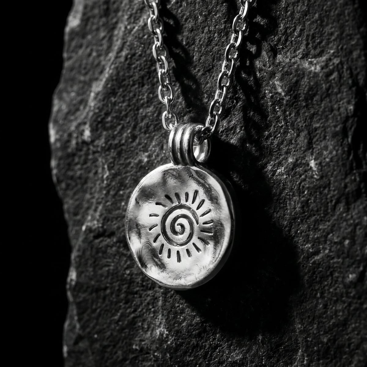 Silver circular pendant with engraved spiral sunburst motif hanging vertically against dark rock.