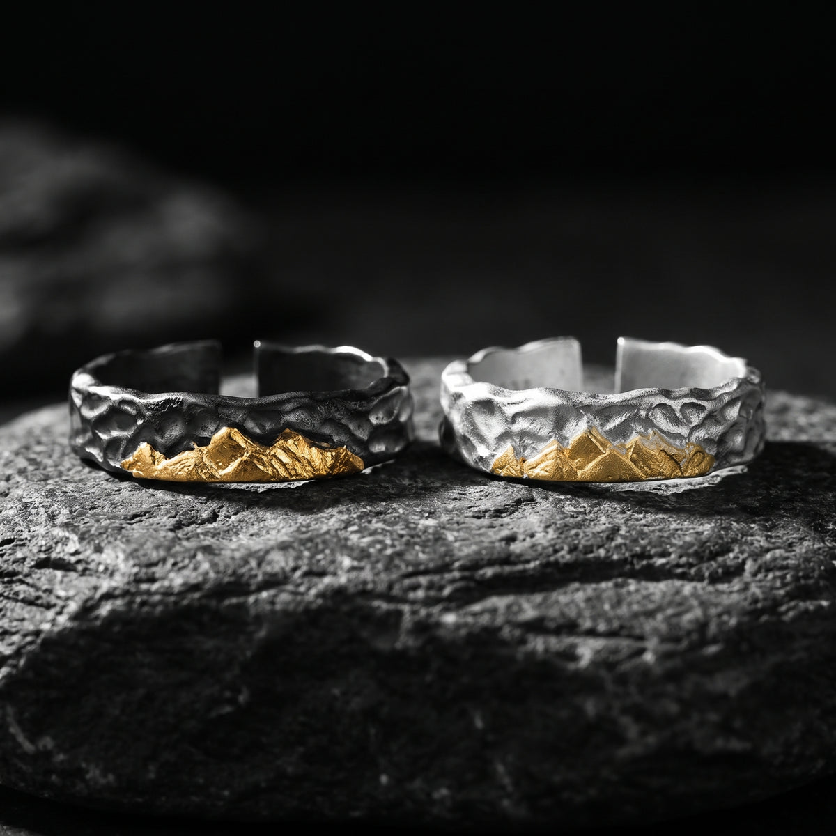 Two open-band rings, one dark-toned and one bright silver, with gold mountain reliefs on a textured rock surface.