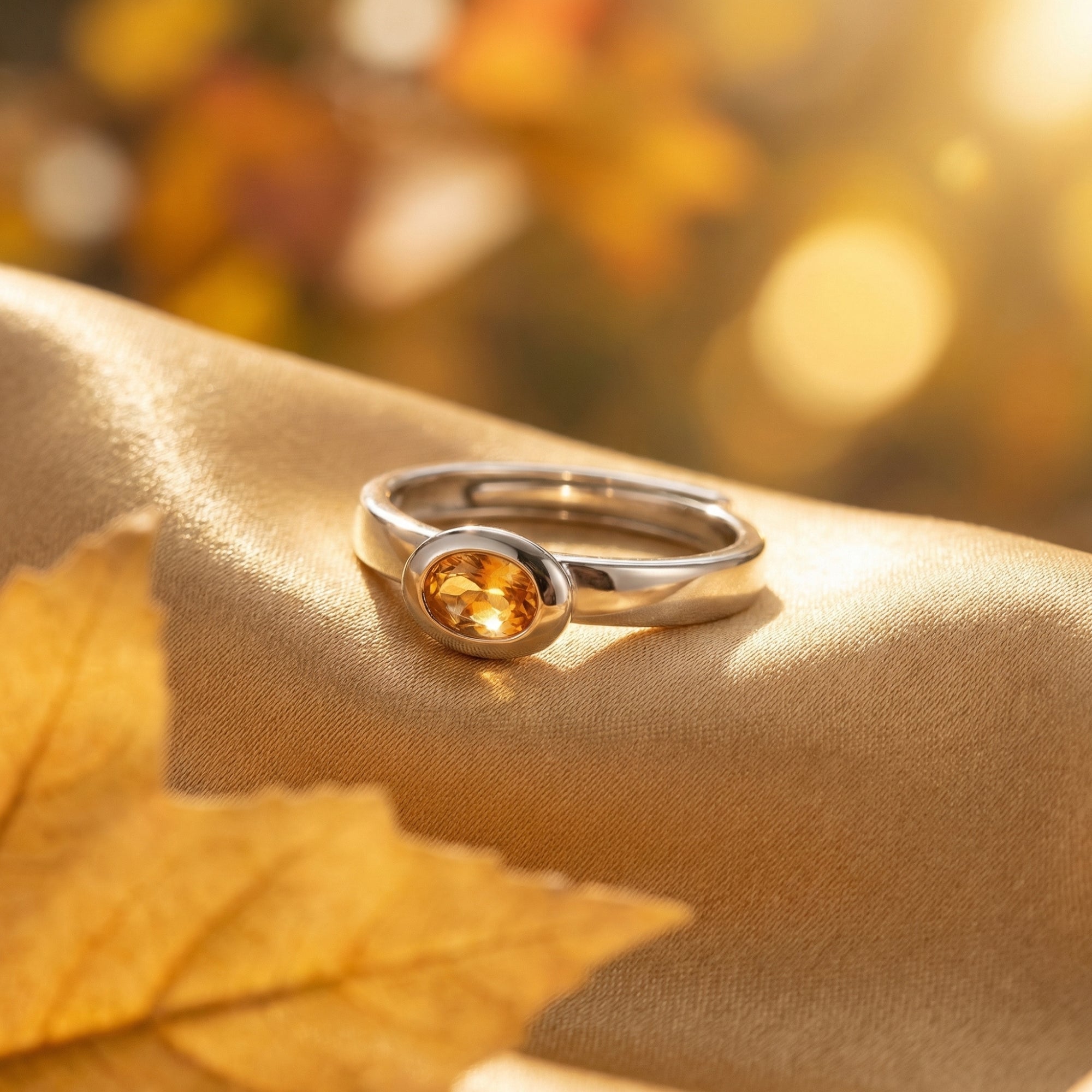 Oval cut natural citrine ring in sterling silver resting on golden fabric with autumn leaves and bokeh background.