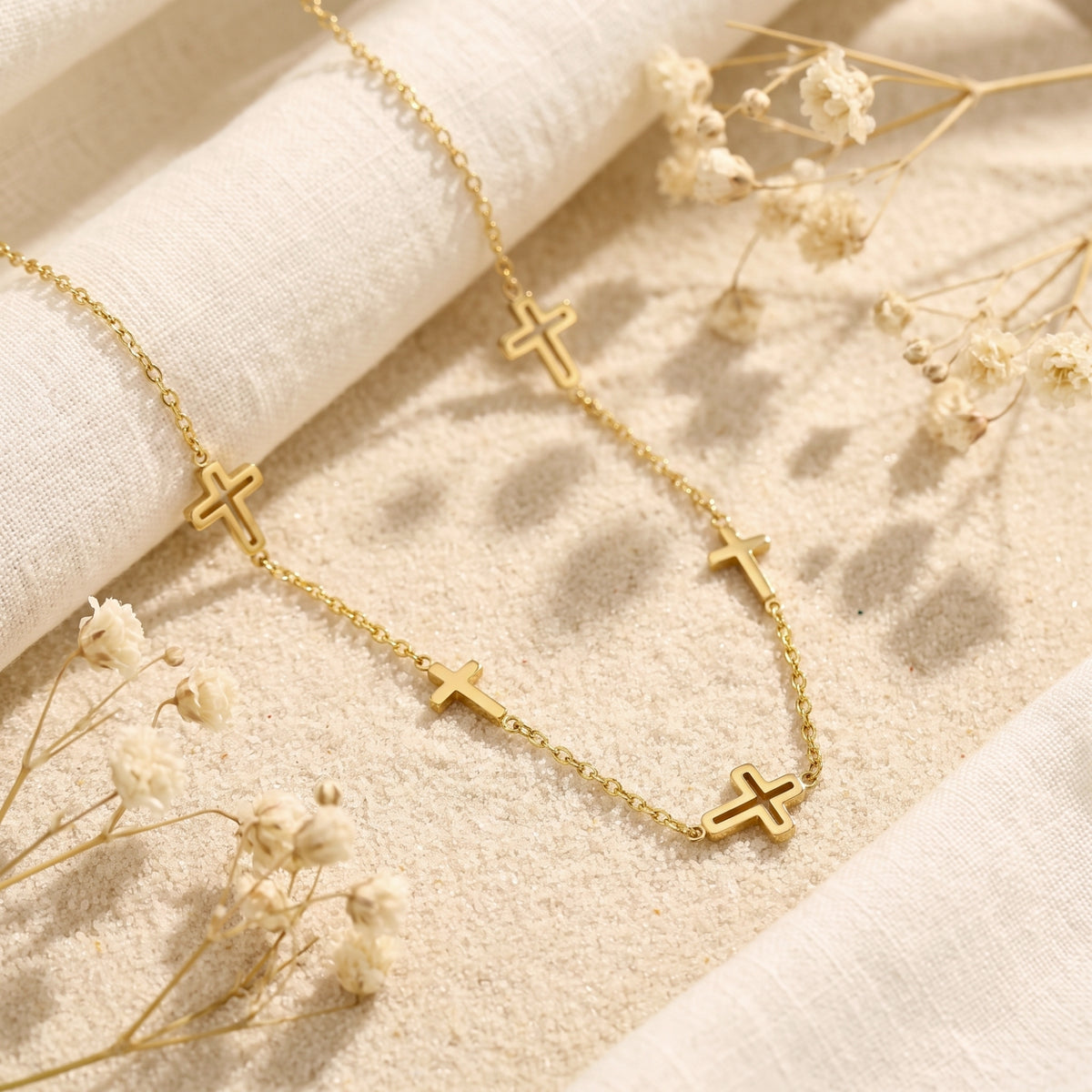 Gold-tone multi-cross station necklace laid on sand with white linen and dried baby's breath flowers.