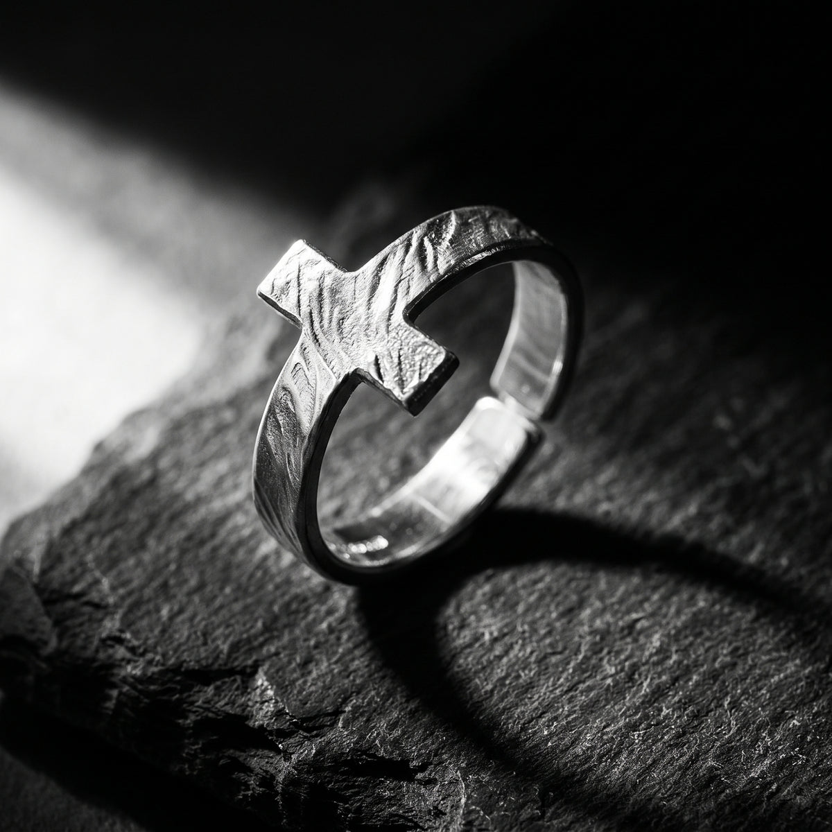 Angled close-up of the textured silver cross ring resting on dark slate rock.