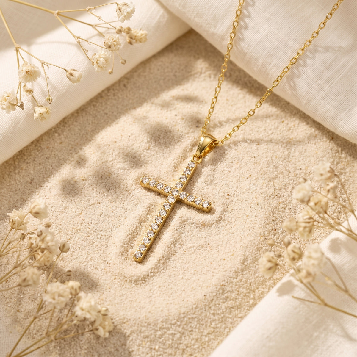 Gold-tone pave cross pendant resting on pale sand, framed by white fabric and dried white flowers.
