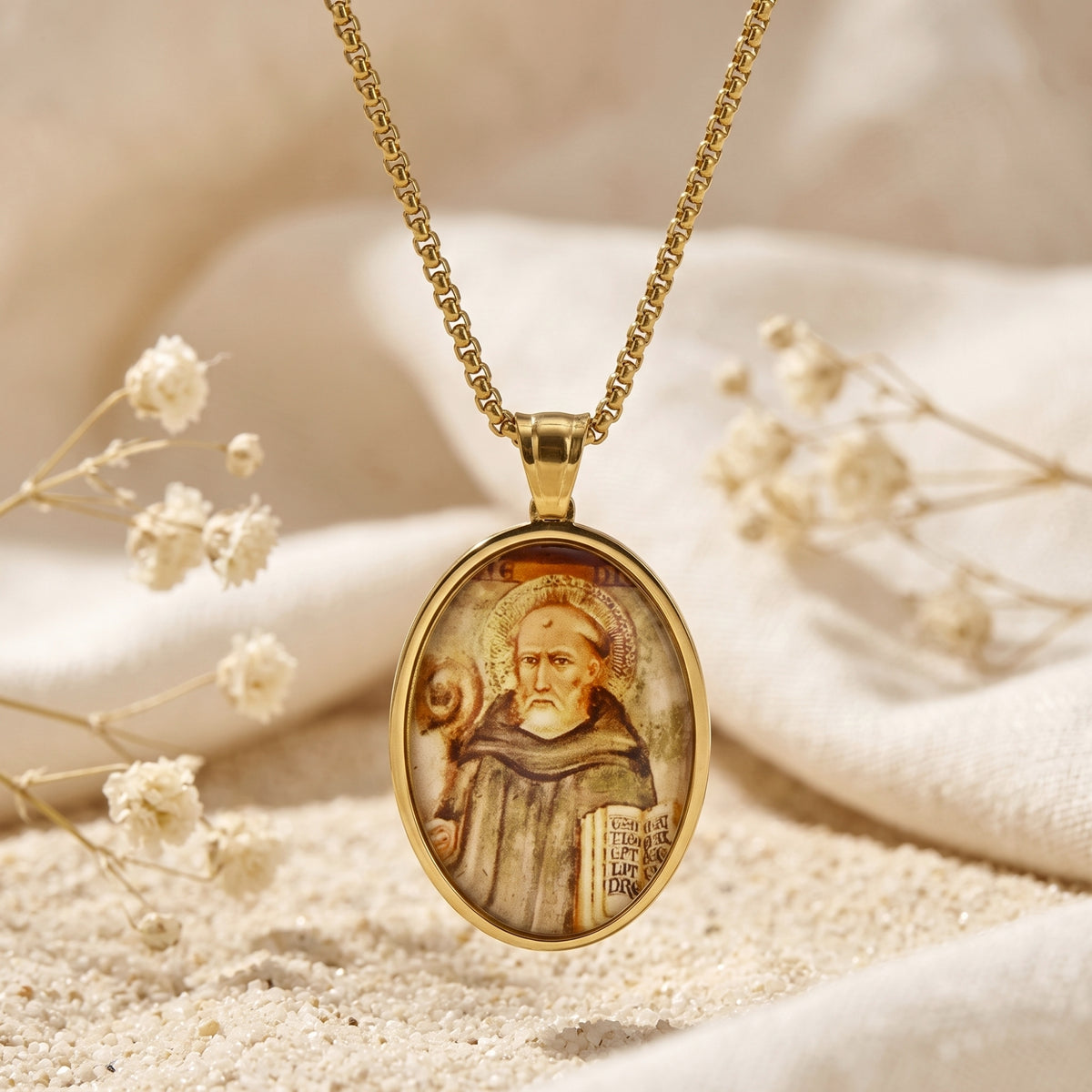 Close-up of a gold-tone oval saint icon pendant necklace hanging over light fabric and sand with dried flowers.