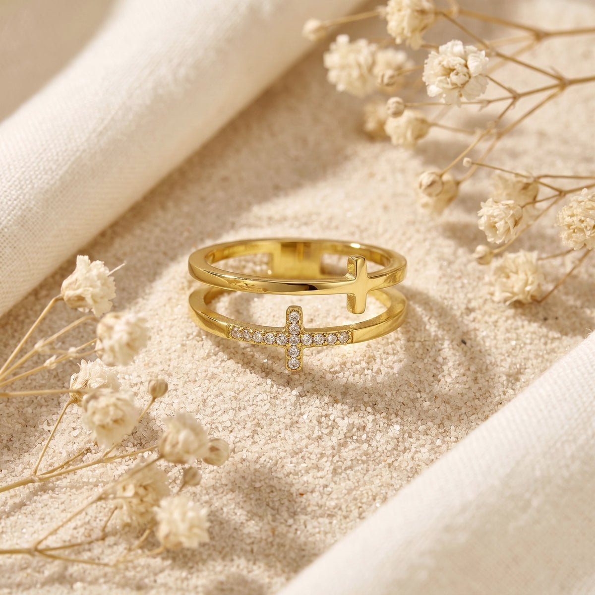 Gold dual-band cross ring on sand, with one polished cross and one crystal-paved cross.