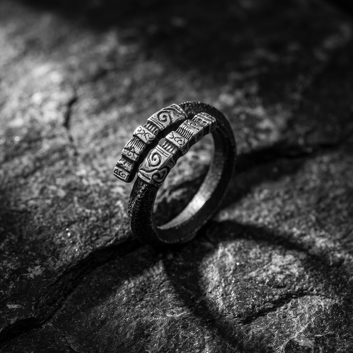 Carved silver open-band ring standing upright on a rough dark stone surface, showing raised relief motifs.