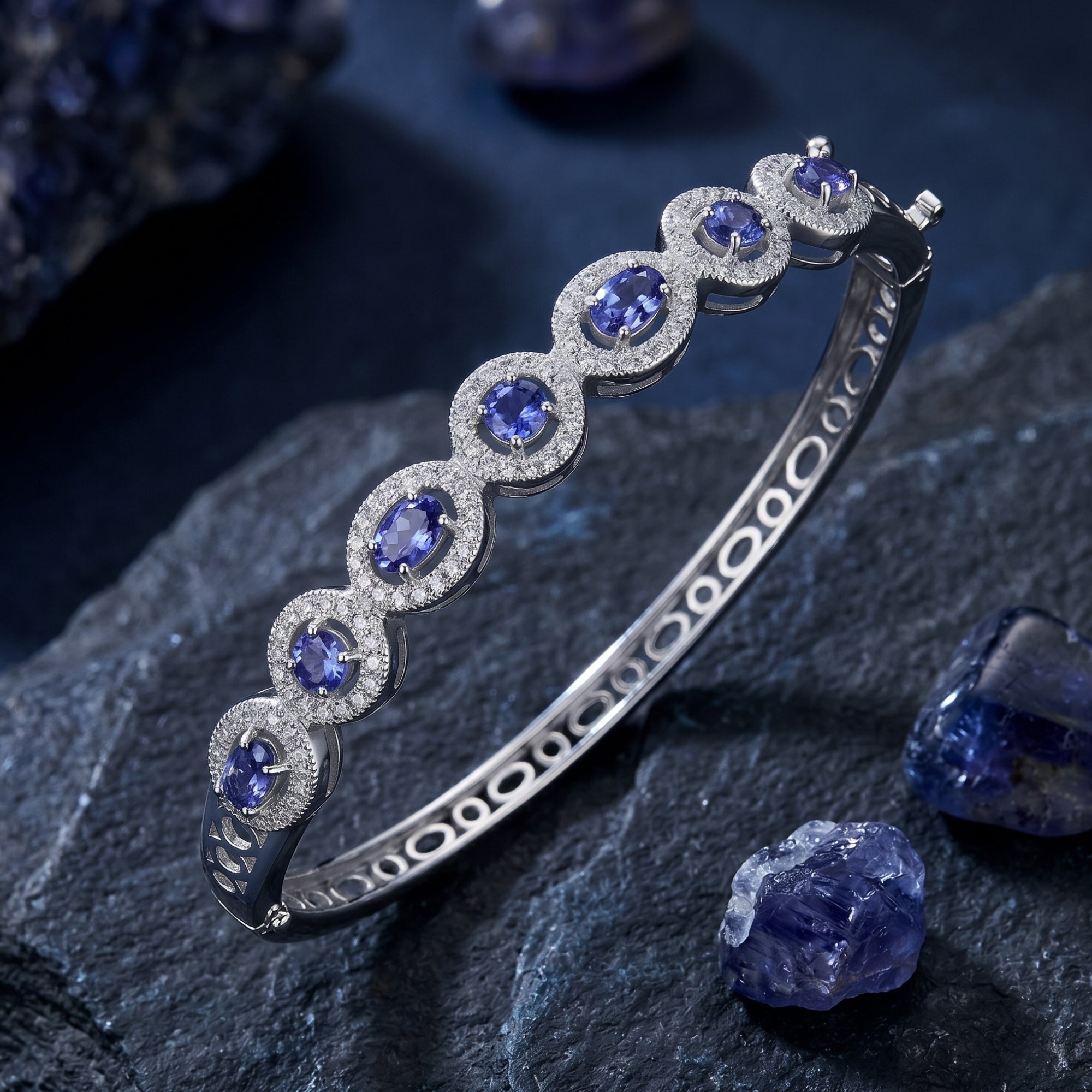 Angled view of a sterling silver tanzanite bracelet with oval and round stones on dark rocks with raw crystals.