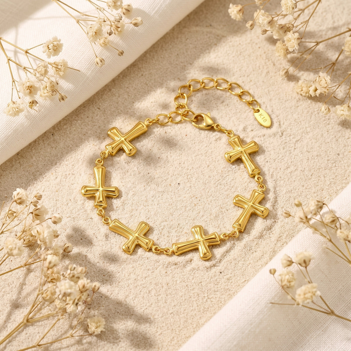 Gold-tone cross station bracelet laid flat on sand, framed by white dried flowers and a fabric edge.