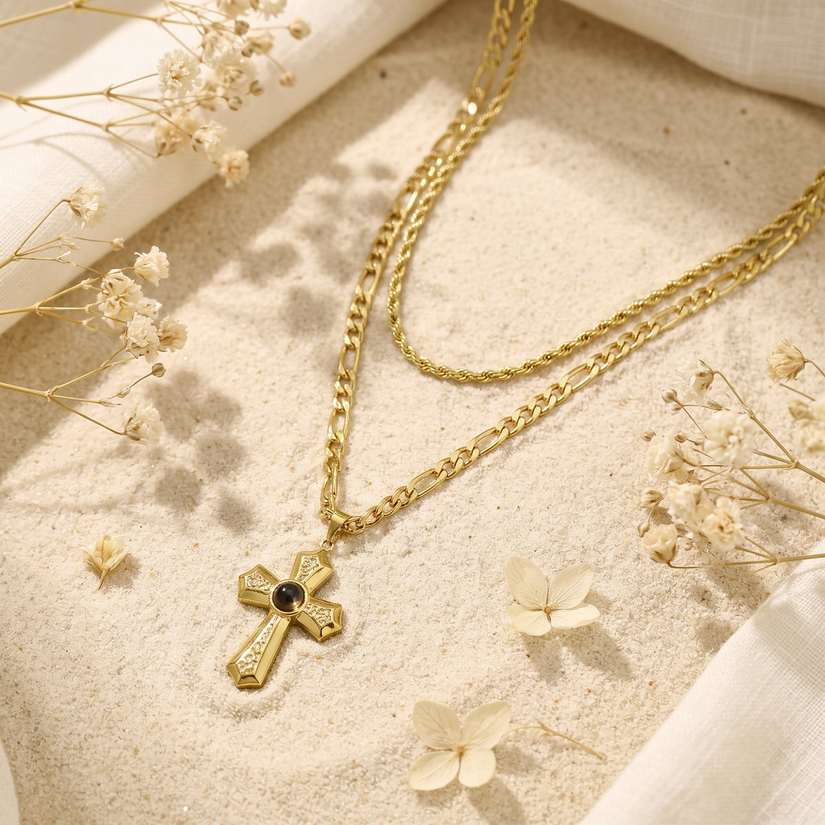 Gold-tone layered cross necklace displayed flat on sand alongside white dried flowers.