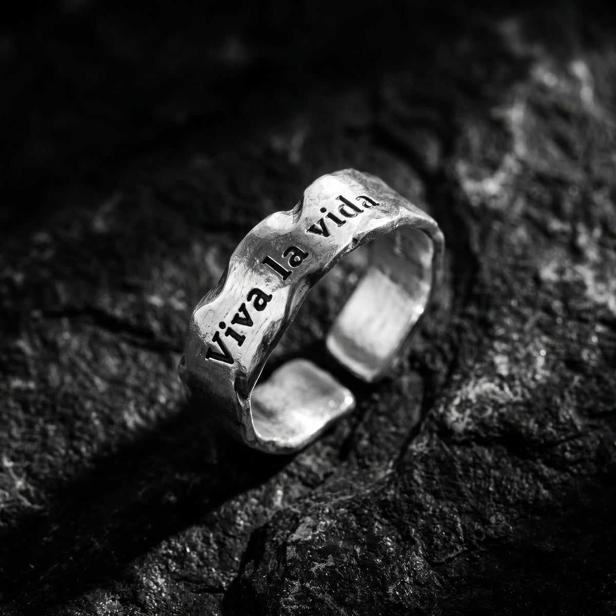 Angled macro view of a silver open-band ring with textured finish and Viva la vida inscription on dark rock.