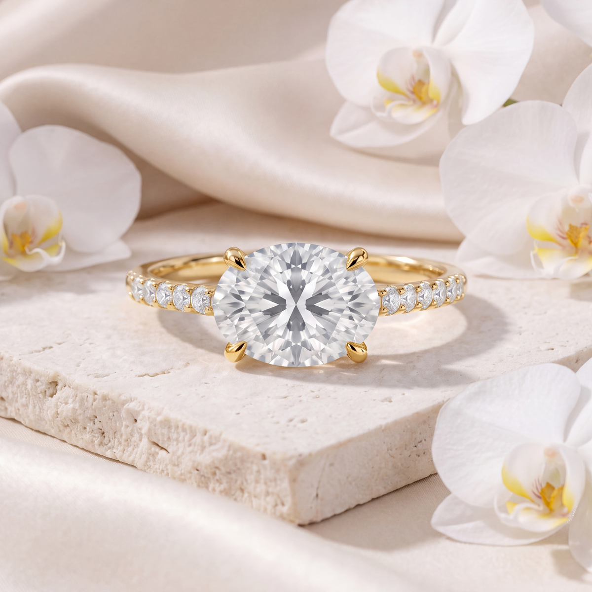 An east-west oval moissanite engagement ring with a pavé band in yellow gold, displayed on travertine stone.