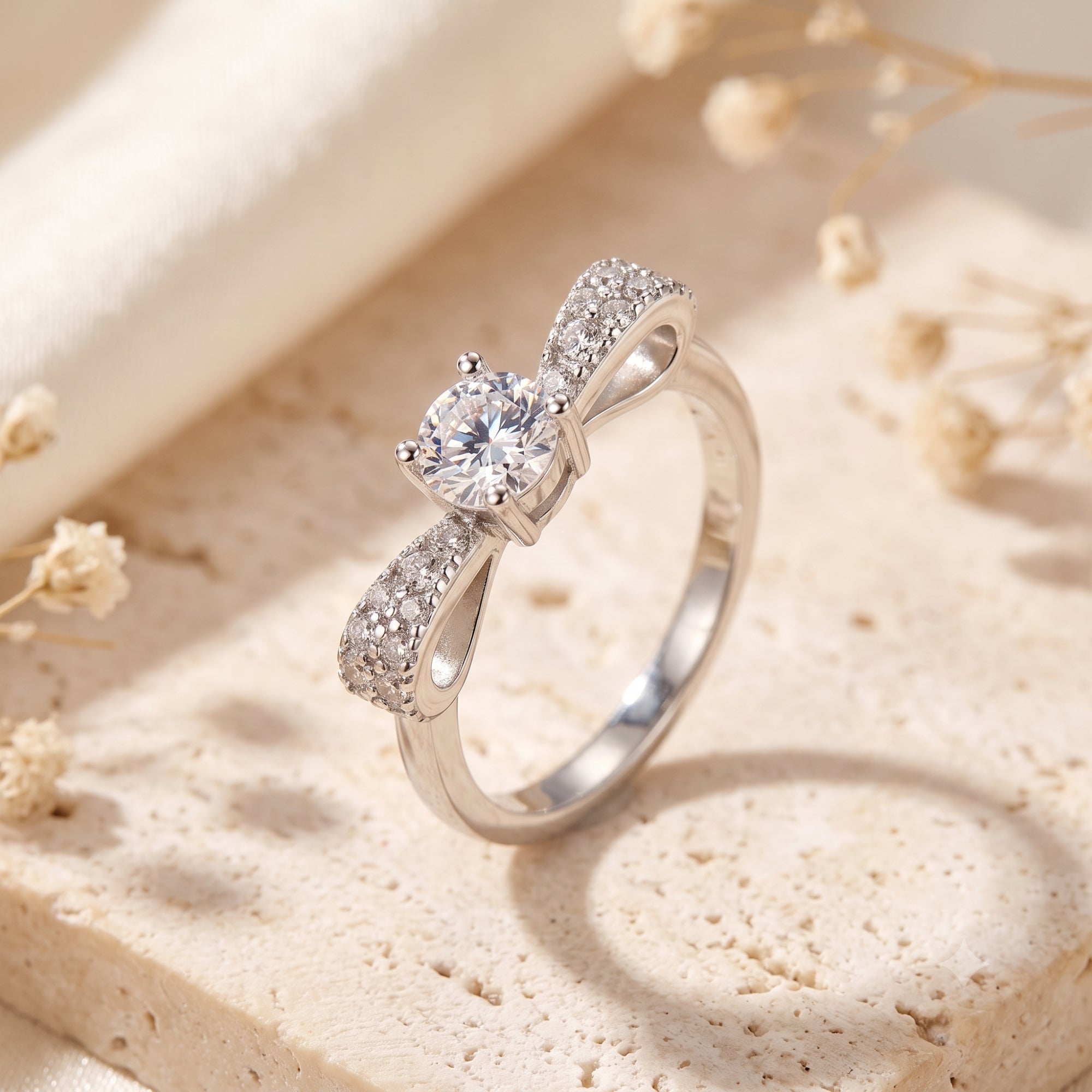 Sterling silver bow promise ring with round cubic zirconia on a stone surface with dried flowers.