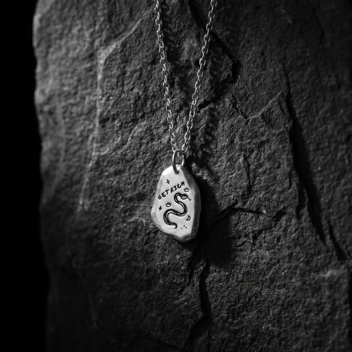 Silver pebble pendant engraved with GET RICH and a snake, hanging against dark slate.