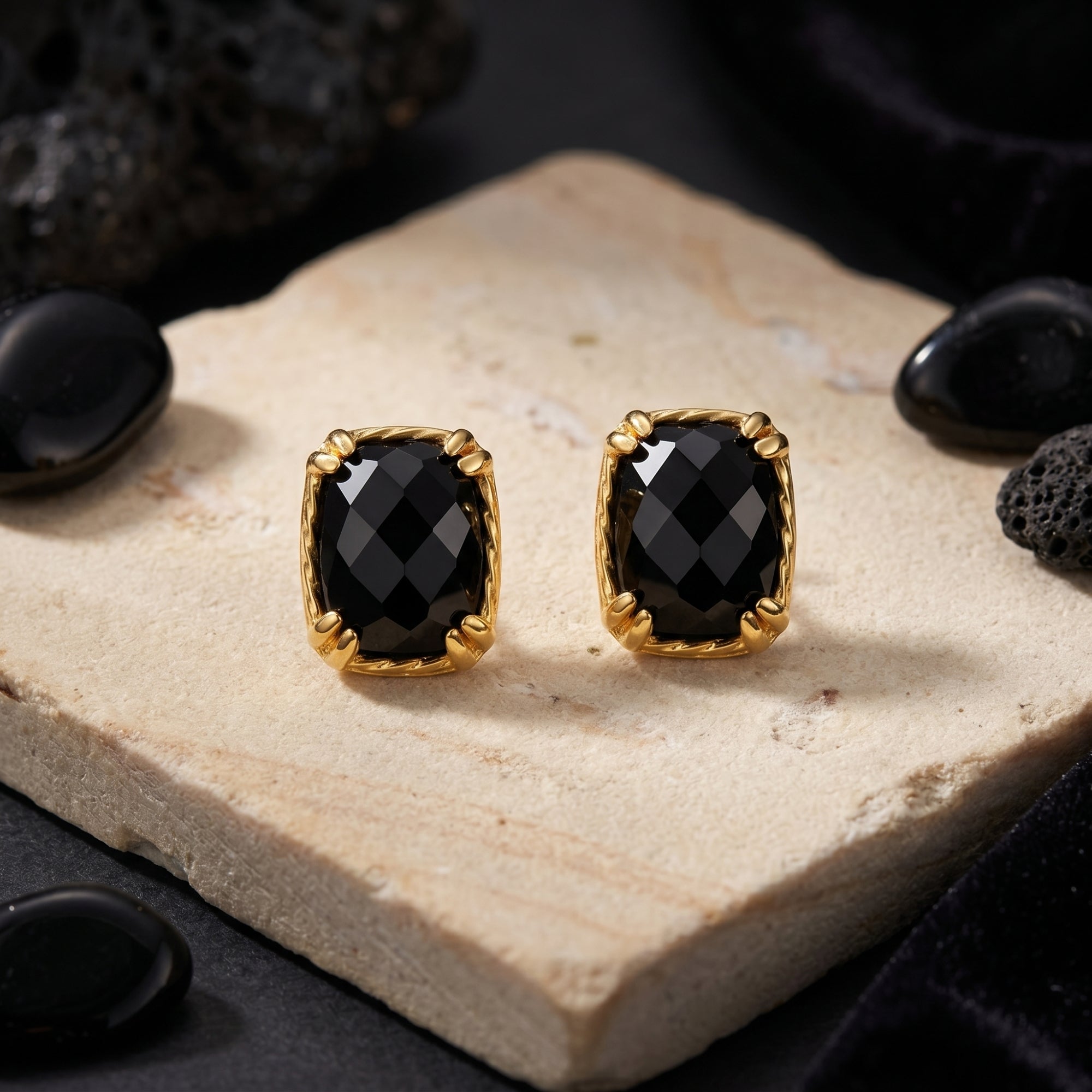 Gold square-cut black onyx stud earrings on a light stone slab with black lava rock props.