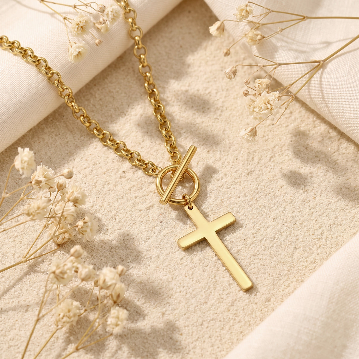 Gold-tone toggle cross necklace resting on fine sand with white fabric and dried flowers.