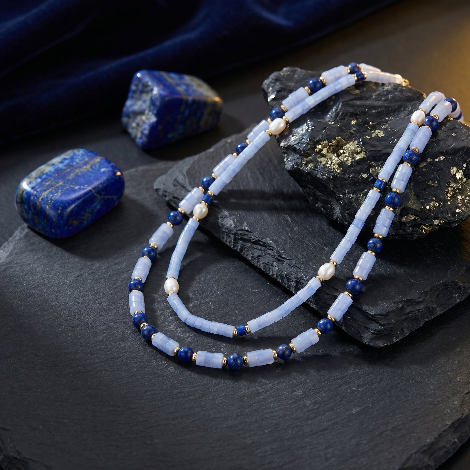 Layered gold plated necklace with cylindrical light blue beads, lapis, and pearls on dark slate with raw stones.