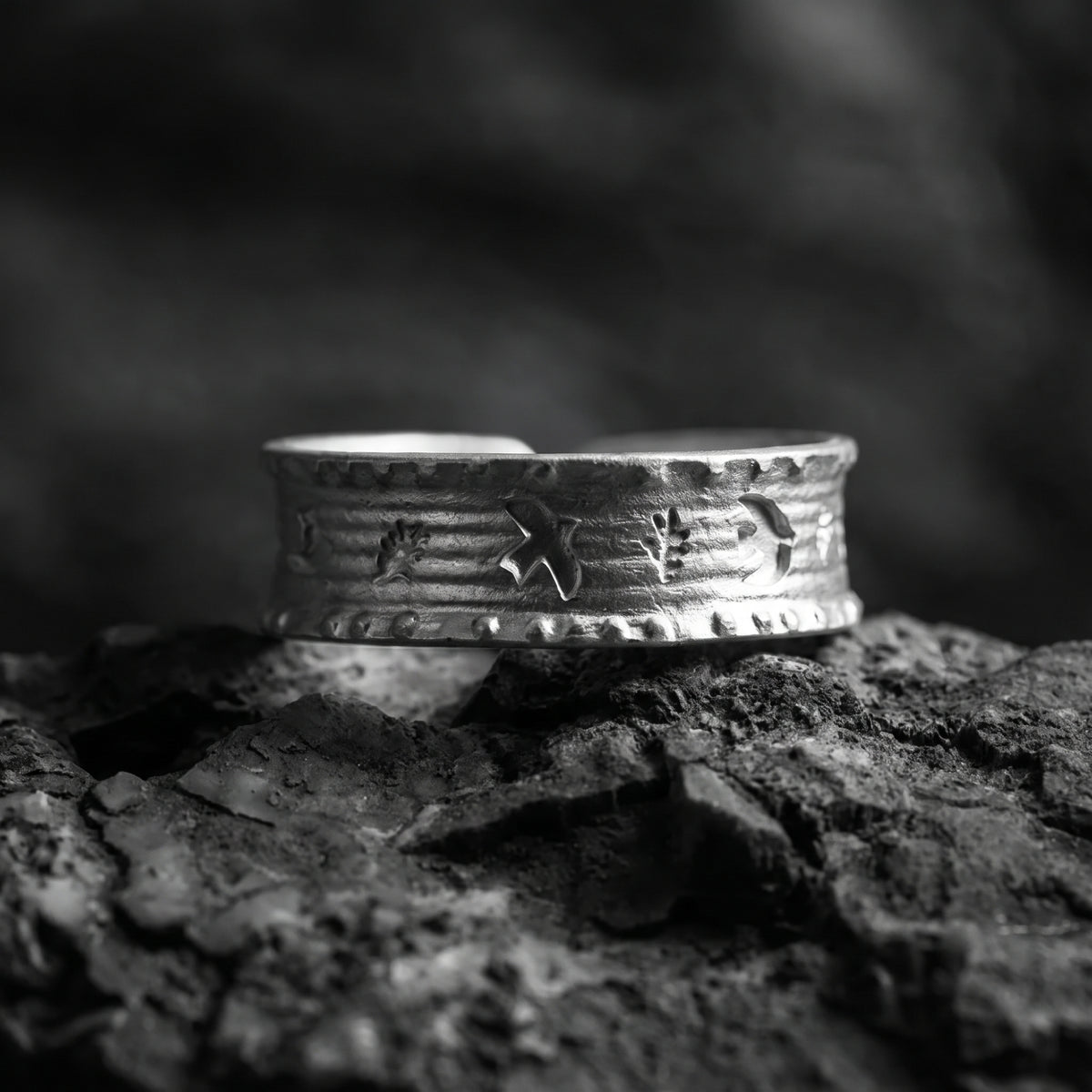 Wide silver ring with raised symbolic motifs and horizontal texture, resting on dark textured rocks.