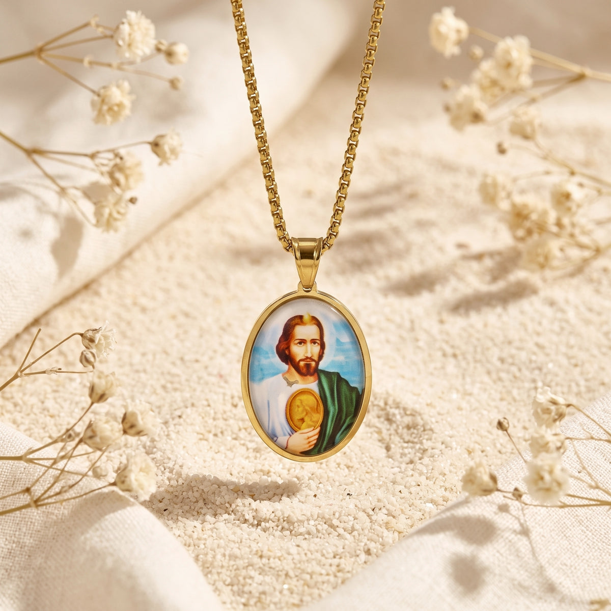 Gold-tone St. Jude medallion pendant hanging vertically on a chain over white sand and dried flowers.
