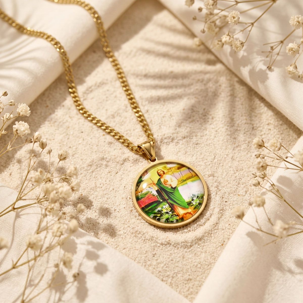 Circular gold-tone St. Jude pendant necklace laid on fine sand with white fabric and flowers.