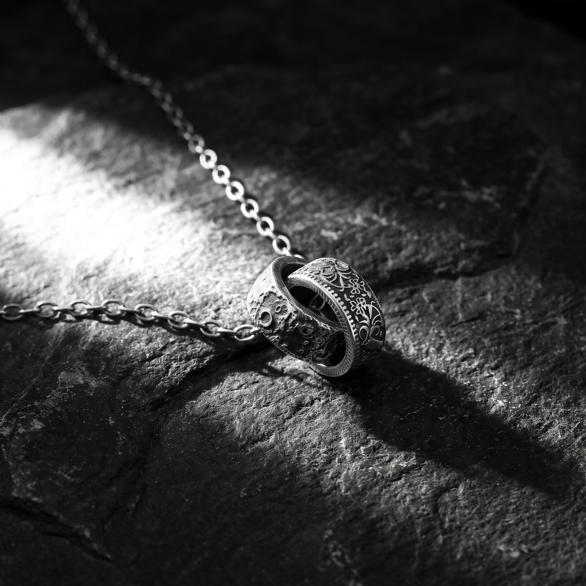Interlocking cratered and engraved silver ring pendant lying on dark textured stone under a spotlight.